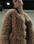 A person wearing the Madison Chubby jacket by Le Superbe in light brown plush faux shearling looks upward in a modern indoor space with exposed ceilings and bright lighting.