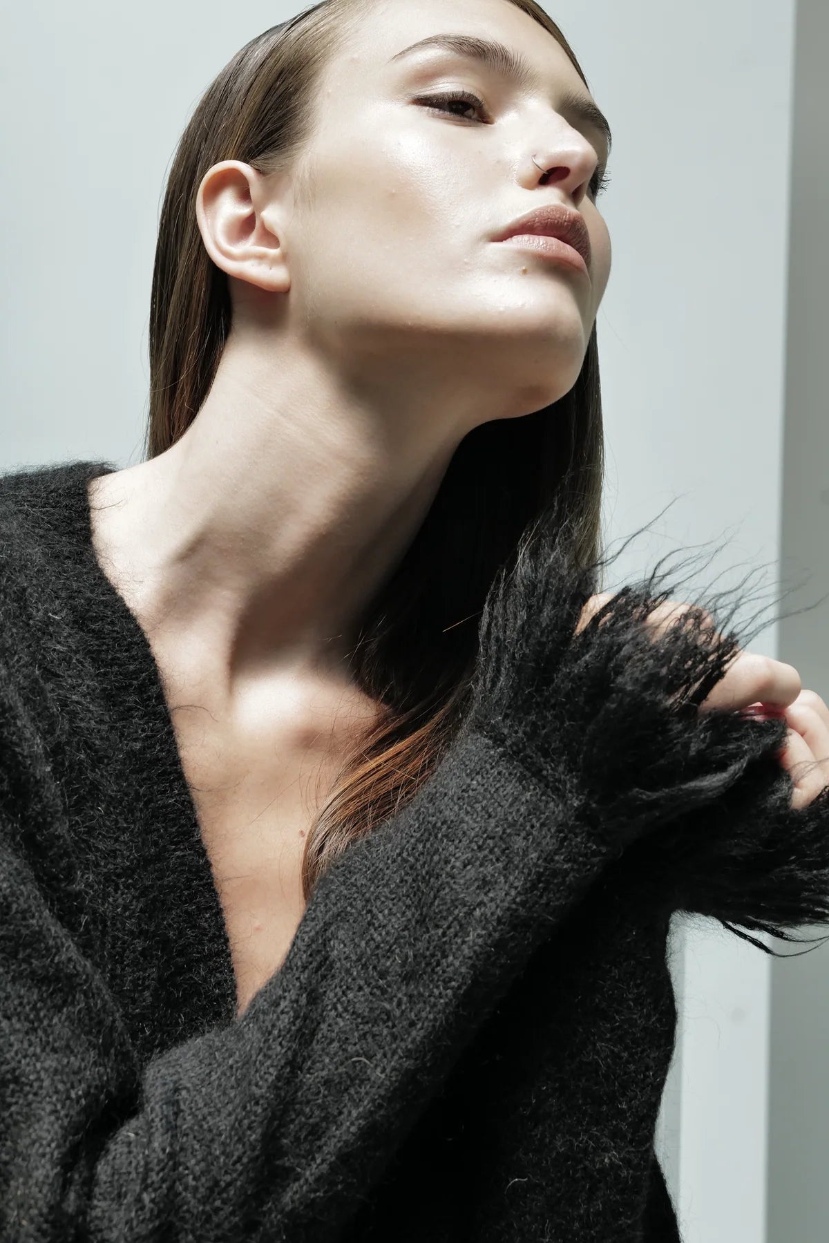 A person with long straight hair wears the Le Superbe Fresange Cardigan, posing with their head tilted back and chin raised. Lighting highlights their smooth skin and the cardigan’s fuzzy knit sleeve detail.