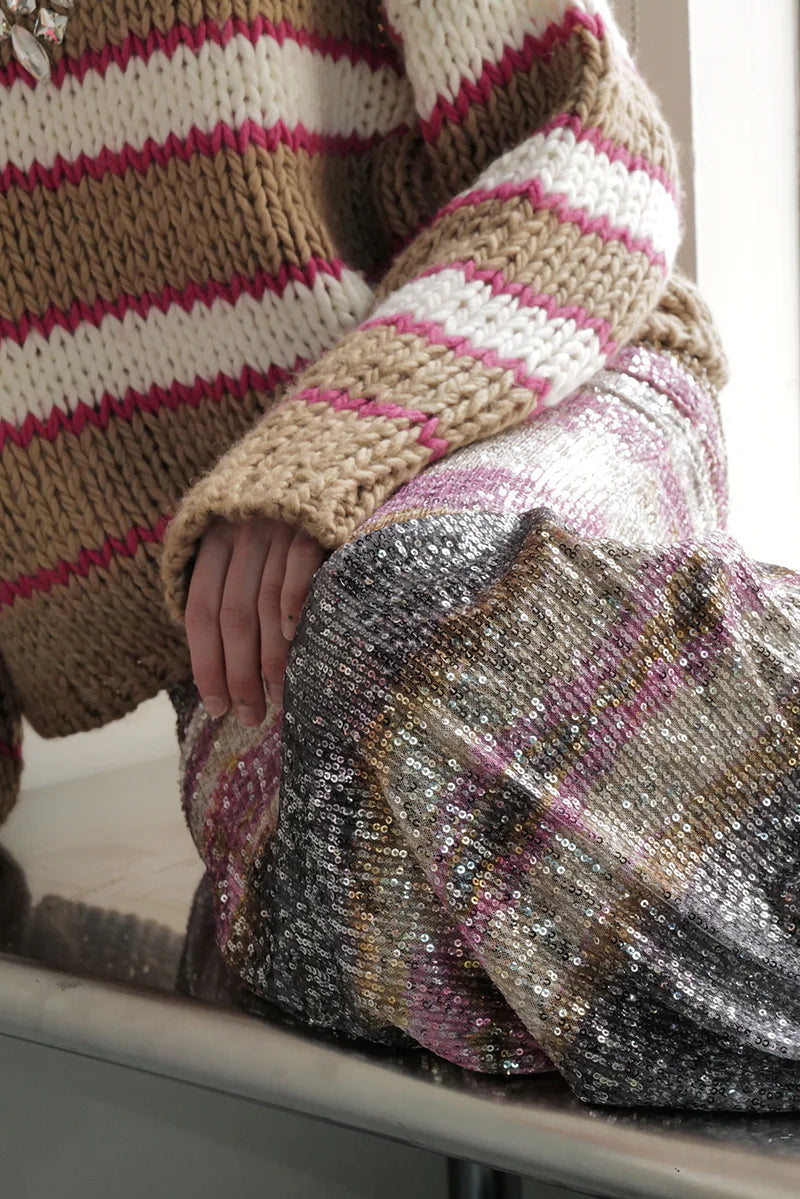 A person sits California cool wearing the Le Superbe Nirvana Sweater—a chunky knit with brown, white, and pink stripes plus a jeweled neckline—one hand on their knee beside a sequined skirt.