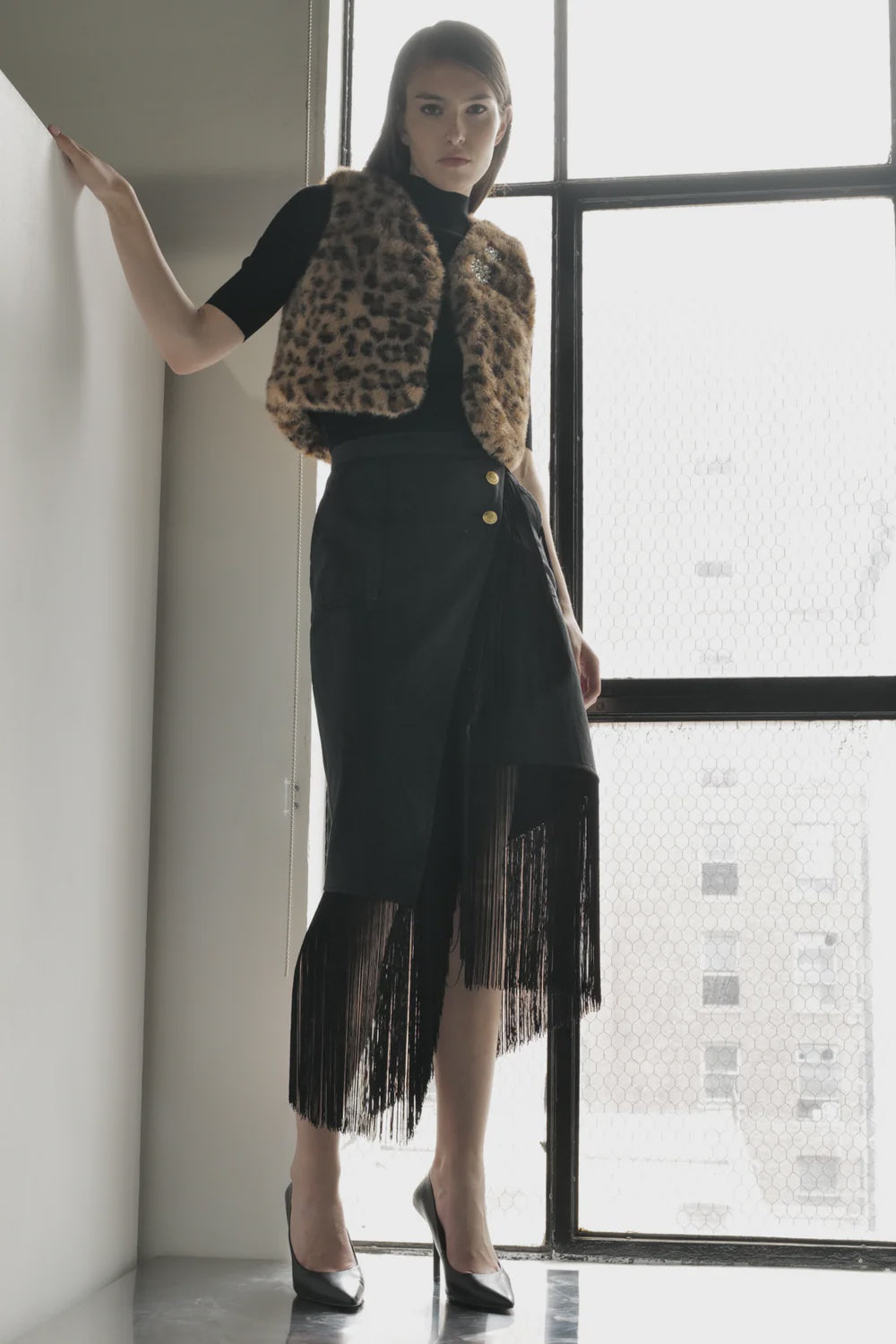 A woman with straight brown hair stands by a large window, confidently wearing the Le Superbe Utility Fringe Skirt, styled with a leopard print vest, black top, and black heels.