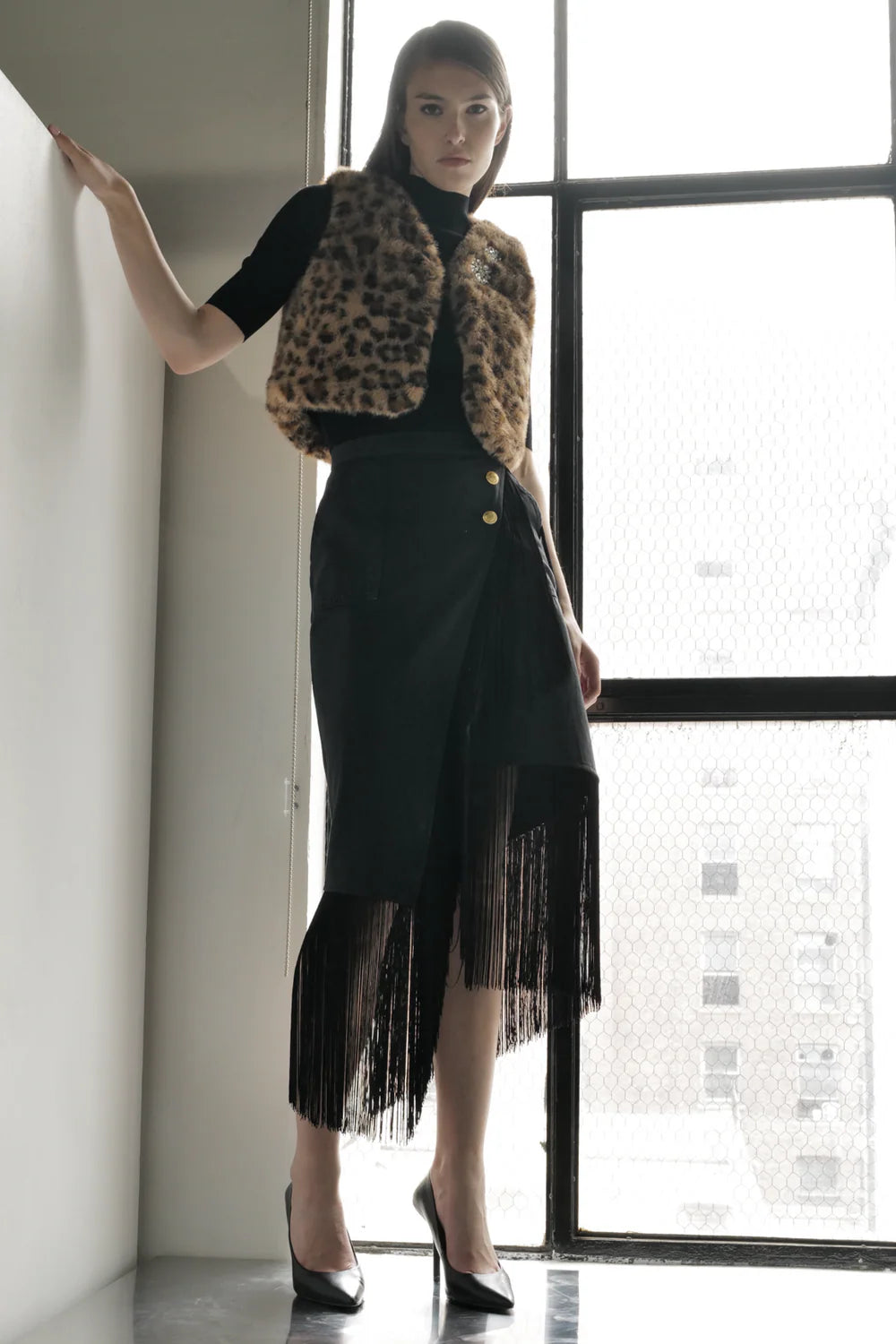 A woman poses indoors by a large window, wearing the Le Superbe Treasure Vest in leopard print over a black top, paired with a fringe-detailed black skirt and black high-heeled shoes.