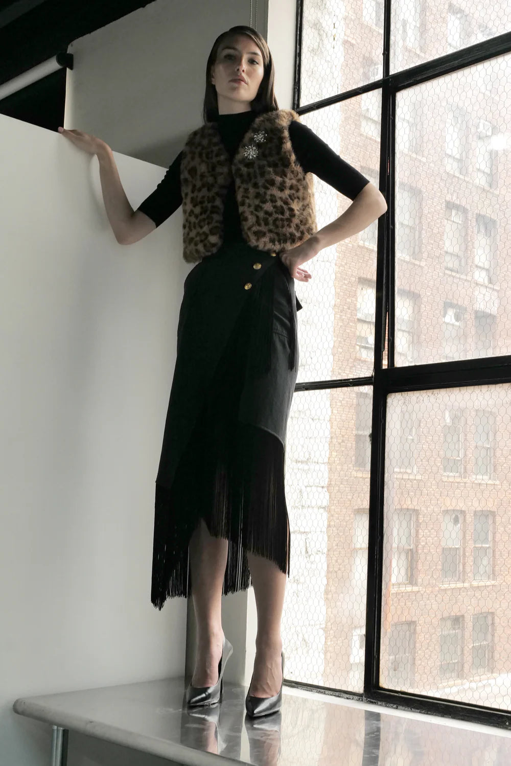 A woman stands by a large window in a leopard print faux fur vest, black top, and the Le Superbe Utility Fringe Skirt with an asymmetrical cut, paired with metallic high heels against a cityscape backdrop.