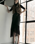 A woman stands by a large window in a leopard print faux fur vest, black top, and the Le Superbe Utility Fringe Skirt with an asymmetrical cut, paired with metallic high heels against a cityscape backdrop.