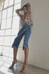 A person poses indoors near large windows, exuding stylish confidence while wearing the Le Superbe Its a Wrap Pant paired with a long-sleeve floral blouse and heeled ankle-strap sandals.