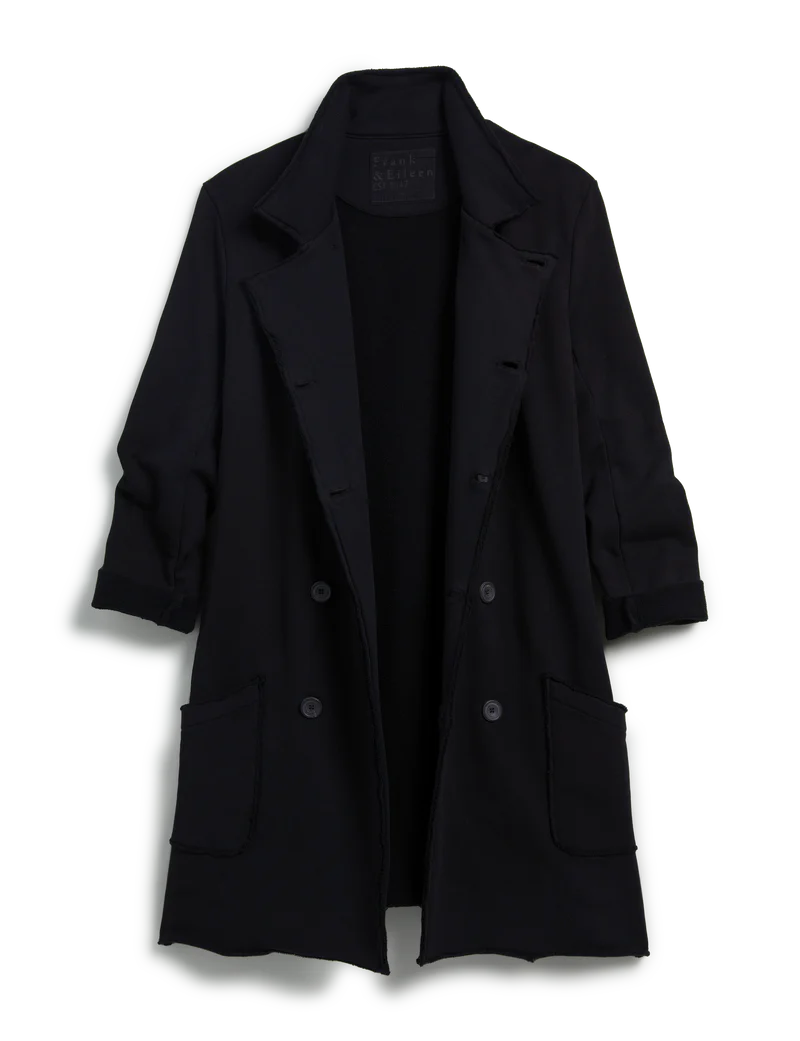 The Frank & Eileen Sweatshirt Trench Coat is a long black, double-breasted English-style coat with notched lapels, large patch pockets, rolled-up sleeves, and is machine-washable. Shown against a plain white background.