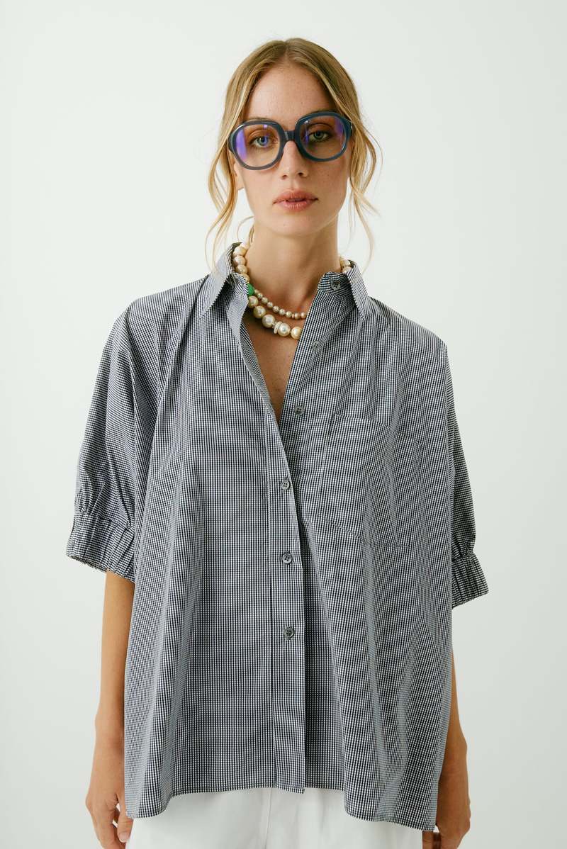 A woman models the Aquarius Cocktail Le Shirt, a cotton oversized button-front shirt dress with short sleeves, styled with layered pearl necklaces and large sunglasses, against a plain light background. Made in Los Angeles.