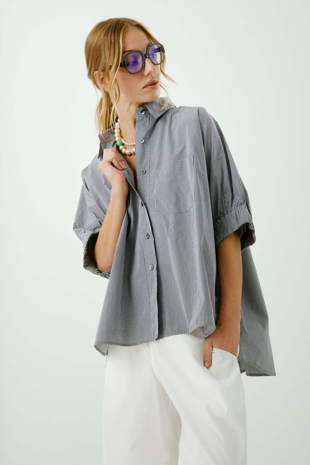 A woman models Aquarius Cocktail's Le Shirt—an oversized, gray pinstriped button-front—styled with blue-tinted sunglasses, white pants, and a chunky beaded necklace, one hand in her pocket and the other holding her collar.