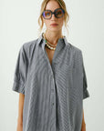 A woman models the Aquarius Cocktail Le Shirt, a cotton oversized button-front shirt dress with short sleeves, styled with layered pearl necklaces and large sunglasses, against a plain light background. Made in Los Angeles.