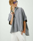 A woman models Aquarius Cocktail's Le Shirt—an oversized, gray pinstriped button-front—styled with blue-tinted sunglasses, white pants, and a chunky beaded necklace, one hand in her pocket and the other holding her collar.