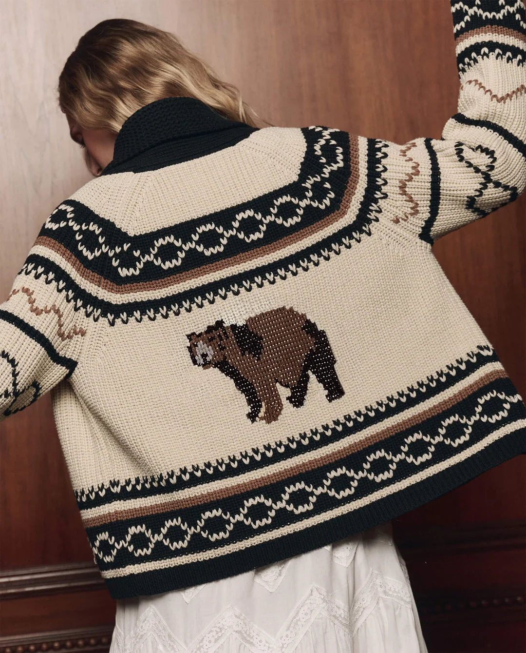The Bear Lodge Cardigan by The Great Inc. in a rustic knit, shown with arms raised against a wooden wall backdrop.