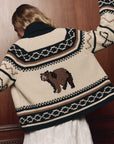 The Bear Lodge Cardigan by The Great Inc. in a rustic knit, shown with arms raised against a wooden wall backdrop.