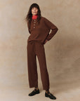 A person with long brown hair stands confidently in a stylish top paired with The Cashmere Lantern Pant from The Great Inc. They have a red scarf around their neck and black shoes on their feet, all set against a neutral, textured background.