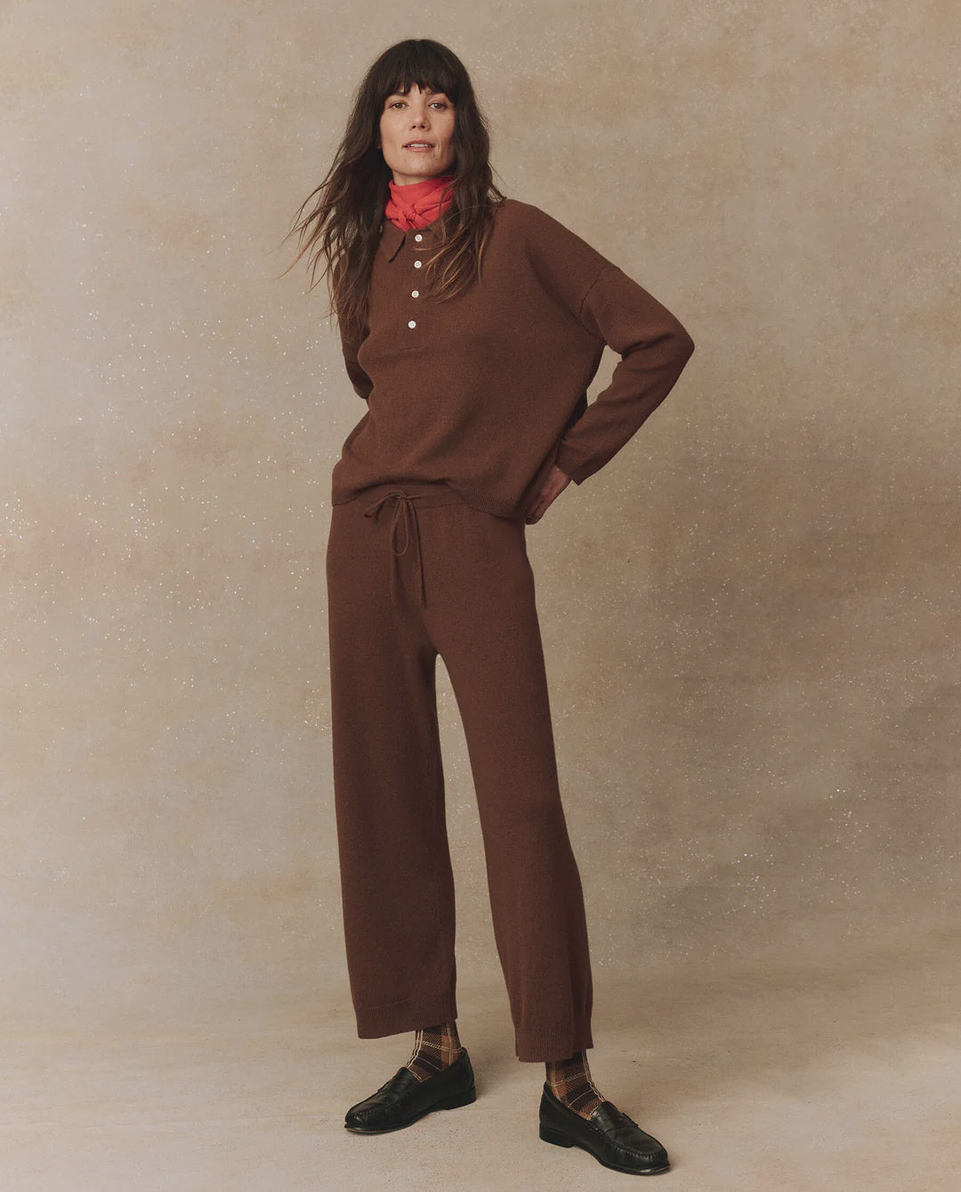 A person with long hair stands against a neutral background, wearing The Great Inc.'s brown long-sleeve top with buttons and The Cashmere Lantern Pant in a matching wide-leg style. A red cashmere scarf complements their look, along with black shoes and plaid socks. They have hands on their hips and are looking at the camera.