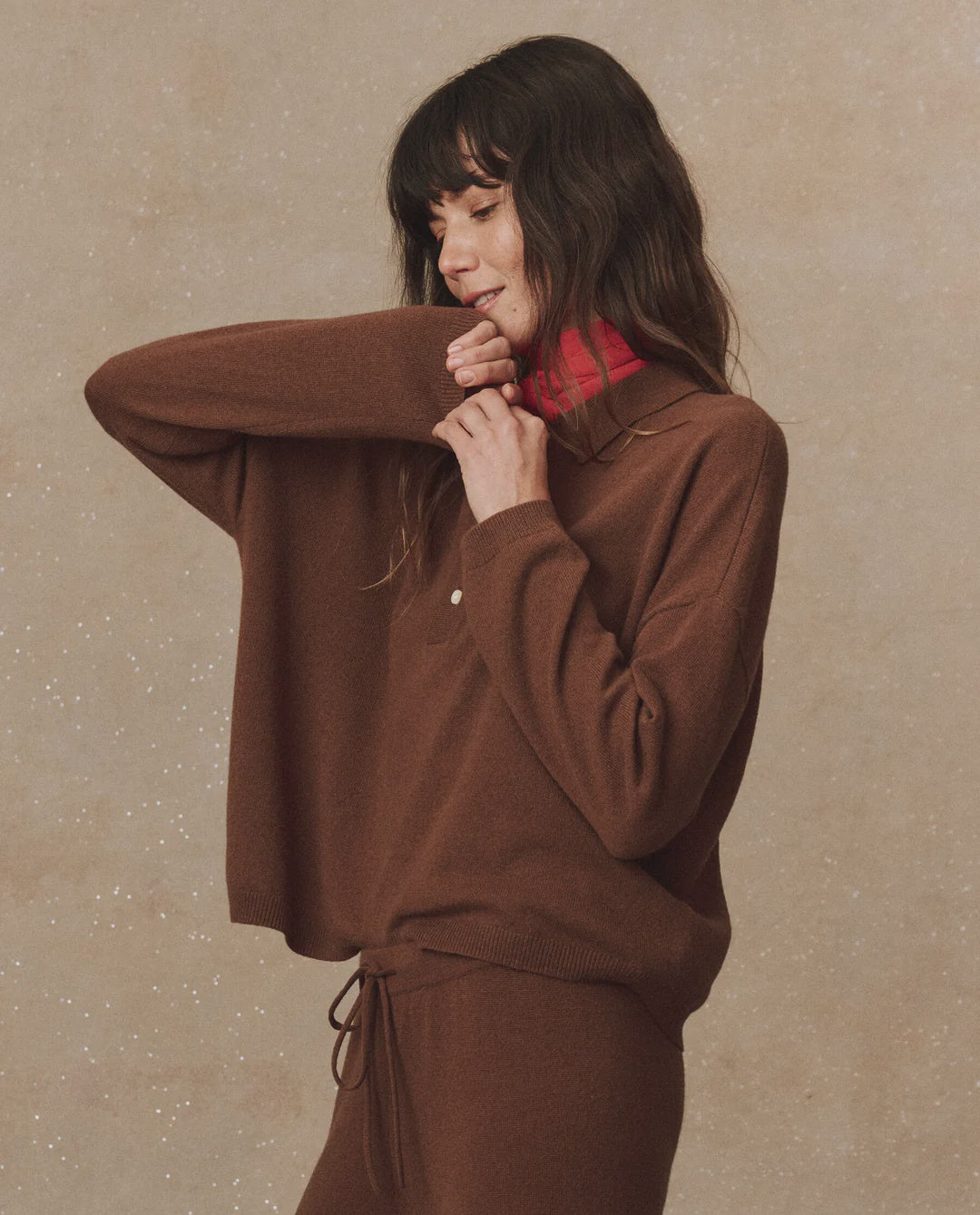 A person with long dark hair smiles while wearing a cashmere polo sweater from The Great Inc., paired with pants. They display a relaxed pose against a neutral, textured background and feature a pink collar, exuding an air reminiscent of vintage menswear.