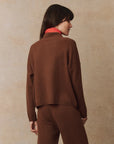 A person with long dark hair stands facing away, dressed in The Great Inc.'s The Cashmere Polo Sweater and matching pants, layered over a red turtleneck. The backdrop features a textured light brown surface with subtle sparkling details.