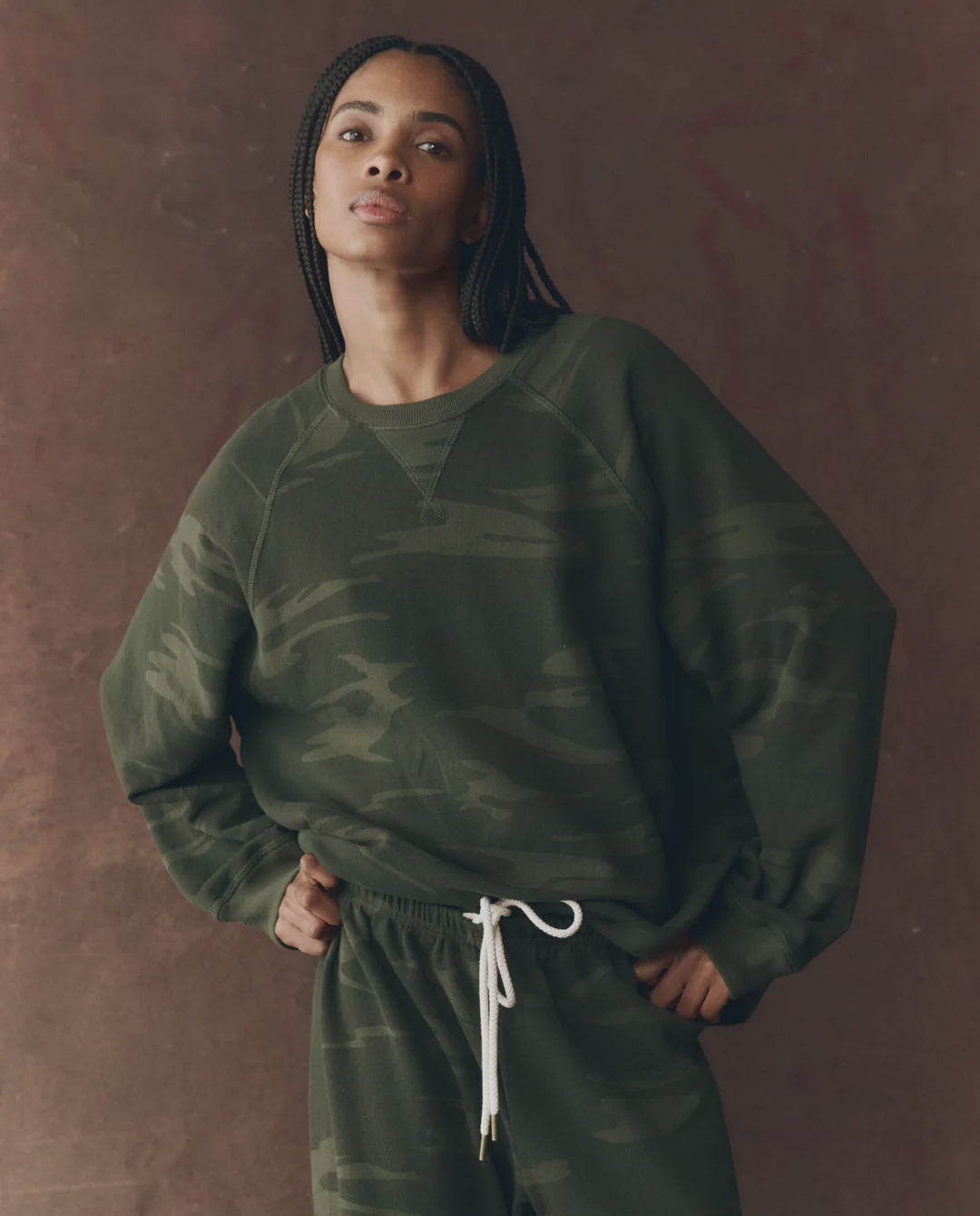 A person with long braids wears The Slouch Sweatshirt by The Great Inc., in dark green camouflage, paired with matching sweatpants with white drawstrings, standing against a brown backdrop and striking a vintage pose.