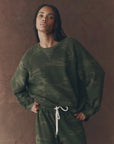 A person with long braids wears The Slouch Sweatshirt by The Great Inc., in dark green camouflage, paired with matching sweatpants with white drawstrings, standing against a brown backdrop and striking a vintage pose.