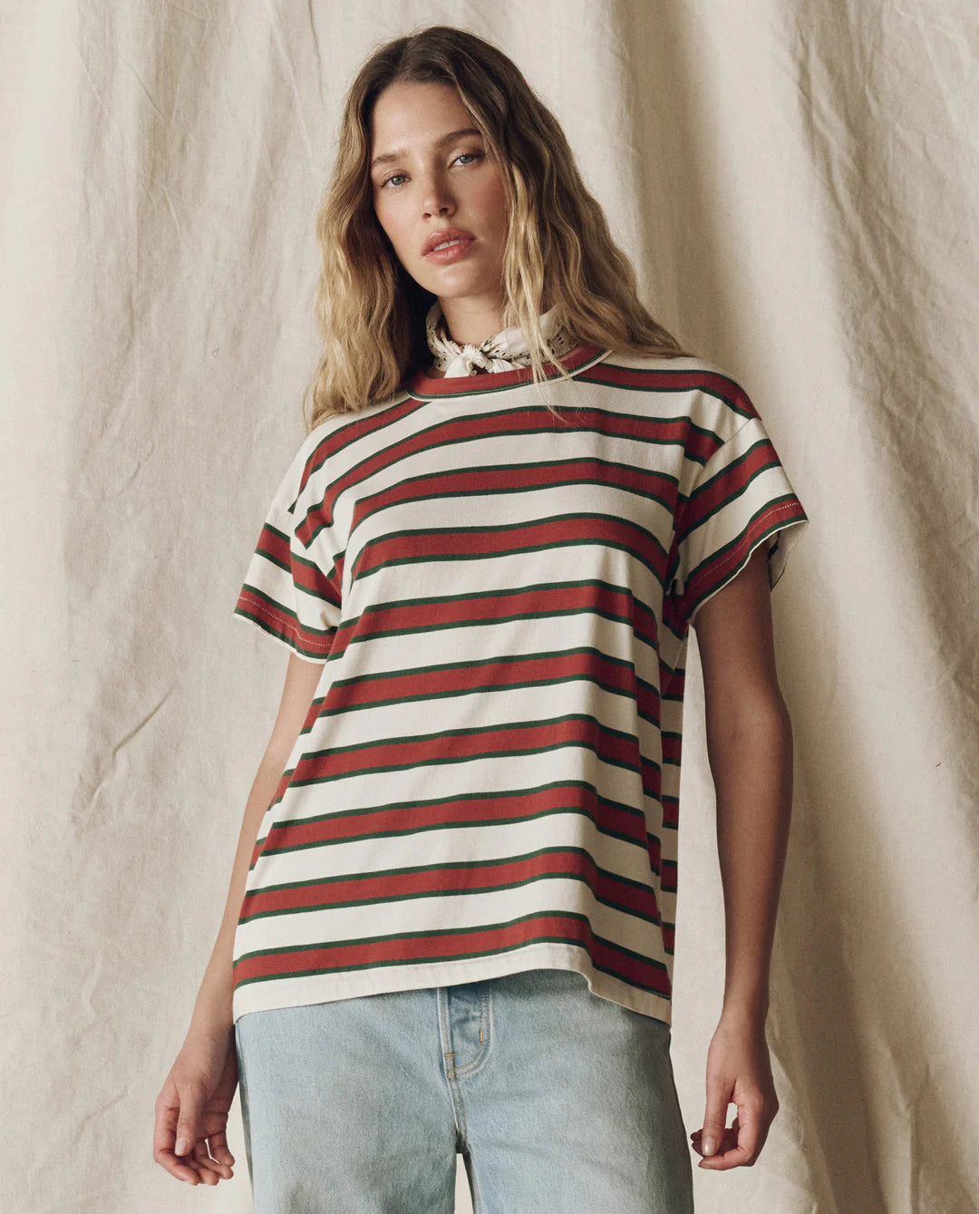 A person with wavy, light brown hair wears The Boxy Crew F25 tee by The Great Inc. with red, green, and white stripes and light blue jeans, standing before a neutral textured fabric backdrop.