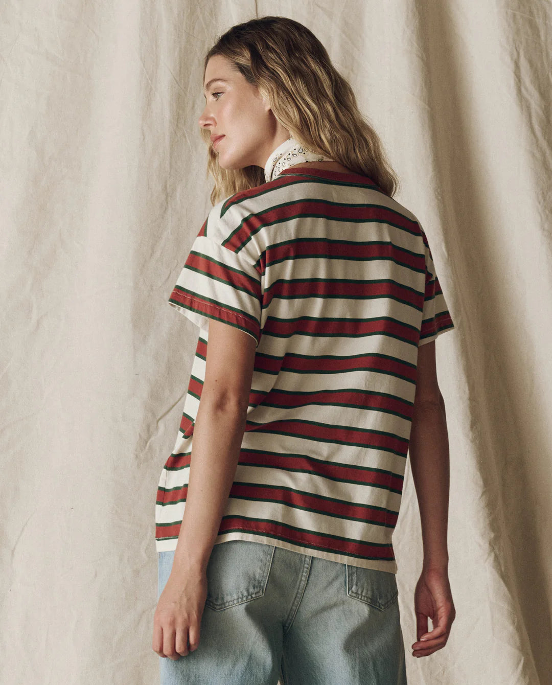 A woman with wavy blonde hair stands with her back to a beige fabric backdrop, wearing The Boxy Crew F25 striped tee by The Great Inc. and light blue jeans.