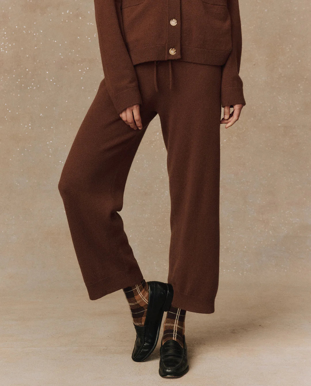 A person is dressed in The Great Inc.'s Cashmere Lantern Pant featuring a wide-leg design in brown, paired with a matching cashmere sweater adorned with large buttons. The ensemble is complemented by plaid socks and black loafers, all set against a speckled beige background.