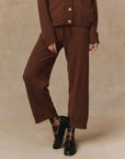 A person is dressed in The Great Inc.'s Cashmere Lantern Pant featuring a wide-leg design in brown, paired with a matching cashmere sweater adorned with large buttons. The ensemble is complemented by plaid socks and black loafers, all set against a speckled beige background.