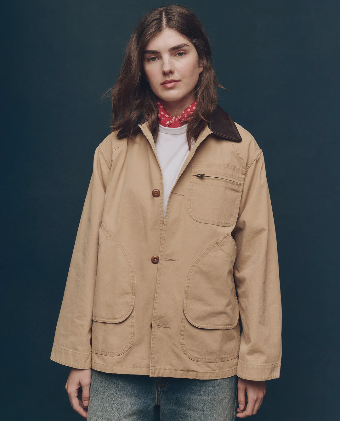 A person with shoulder-length brown hair wears The Great Inc.'s The Barn Jacket—a tan garment-dyed cotton canvas workwear jacket with a leather collar—over a white shirt, blue jeans, and a red scarf, standing against a plain dark background.