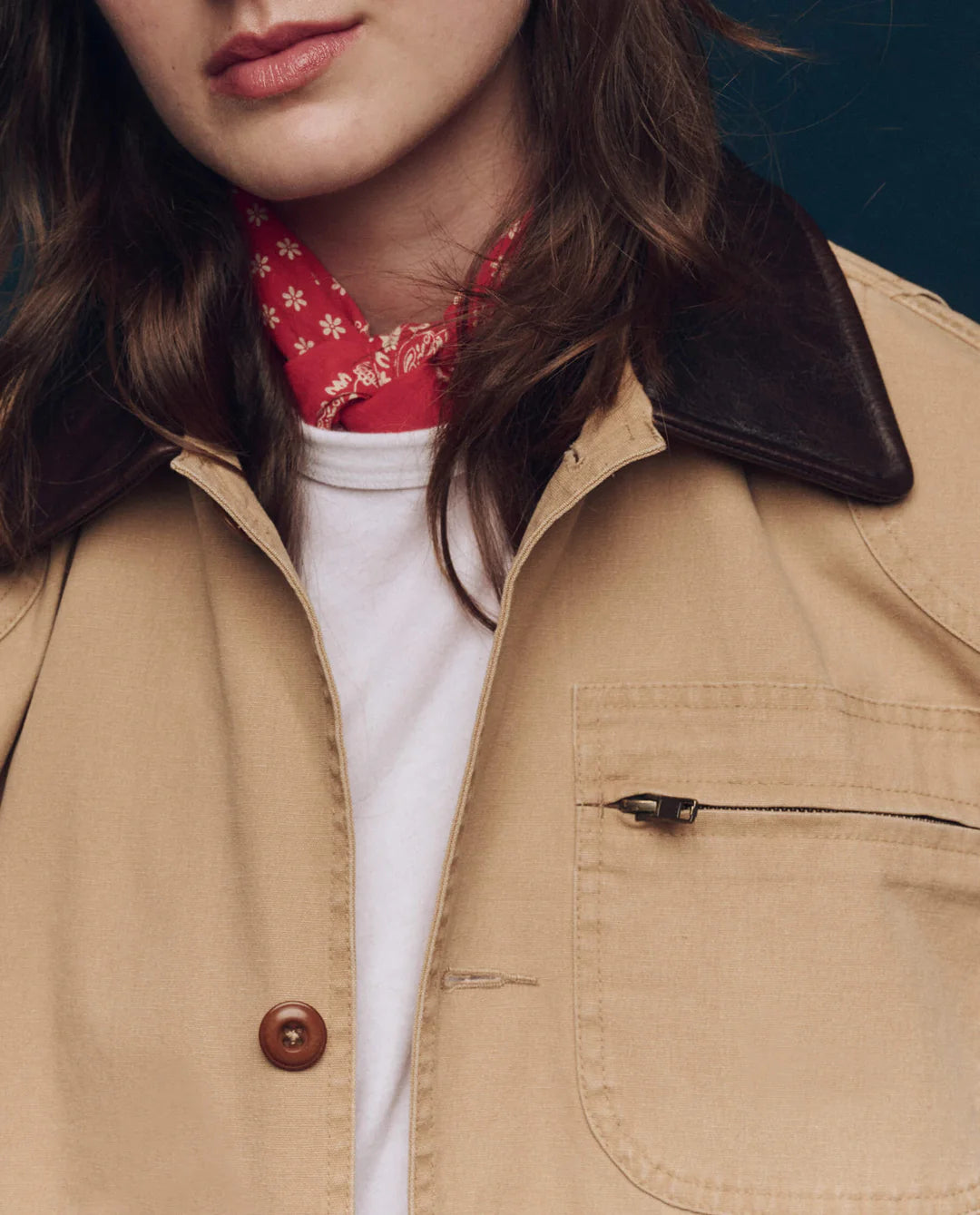 A person wears The Great Inc. Barn Jacket, a tan garment-dyed cotton workwear piece with a dark brown collar, over a white shirt and red patterned bandana. Only the lower half of their face and upper torso are visible.