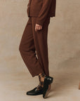 A person is seen wearing The Great Inc.'s Cashmere Lantern Pant along with a matching top, set against a speckled beige background. They pair the outfit with black loafers and brown plaid socks, crossing one foot over the other, with the photo highlighting the lower half of their body.