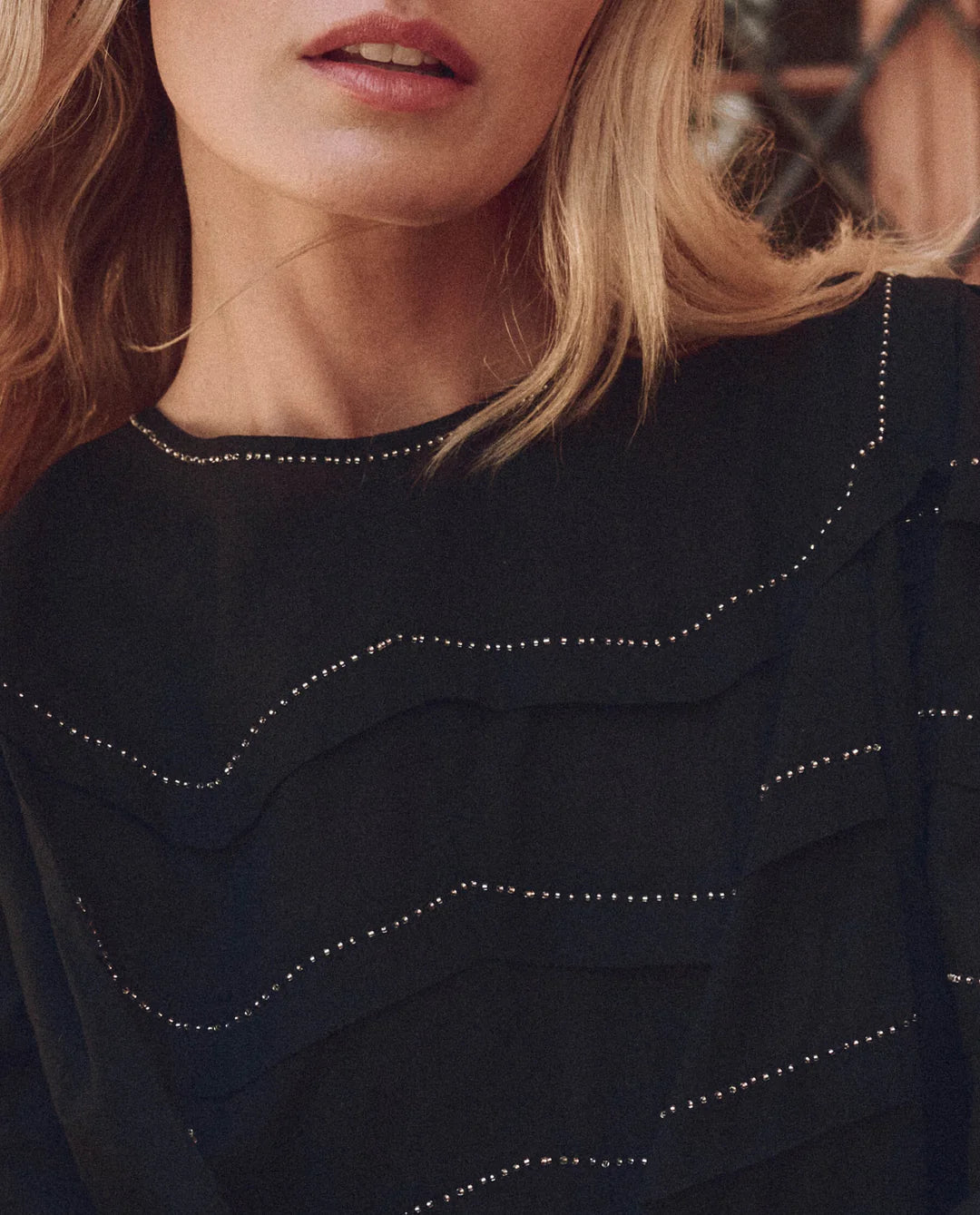 A close-up of a person with long blond hair wearing The Beaded Chevron Top by The Great Inc. highlights their lips and upper torso, showcasing the textured detail and vintage-inspired beadwork that exudes timeless elegance.