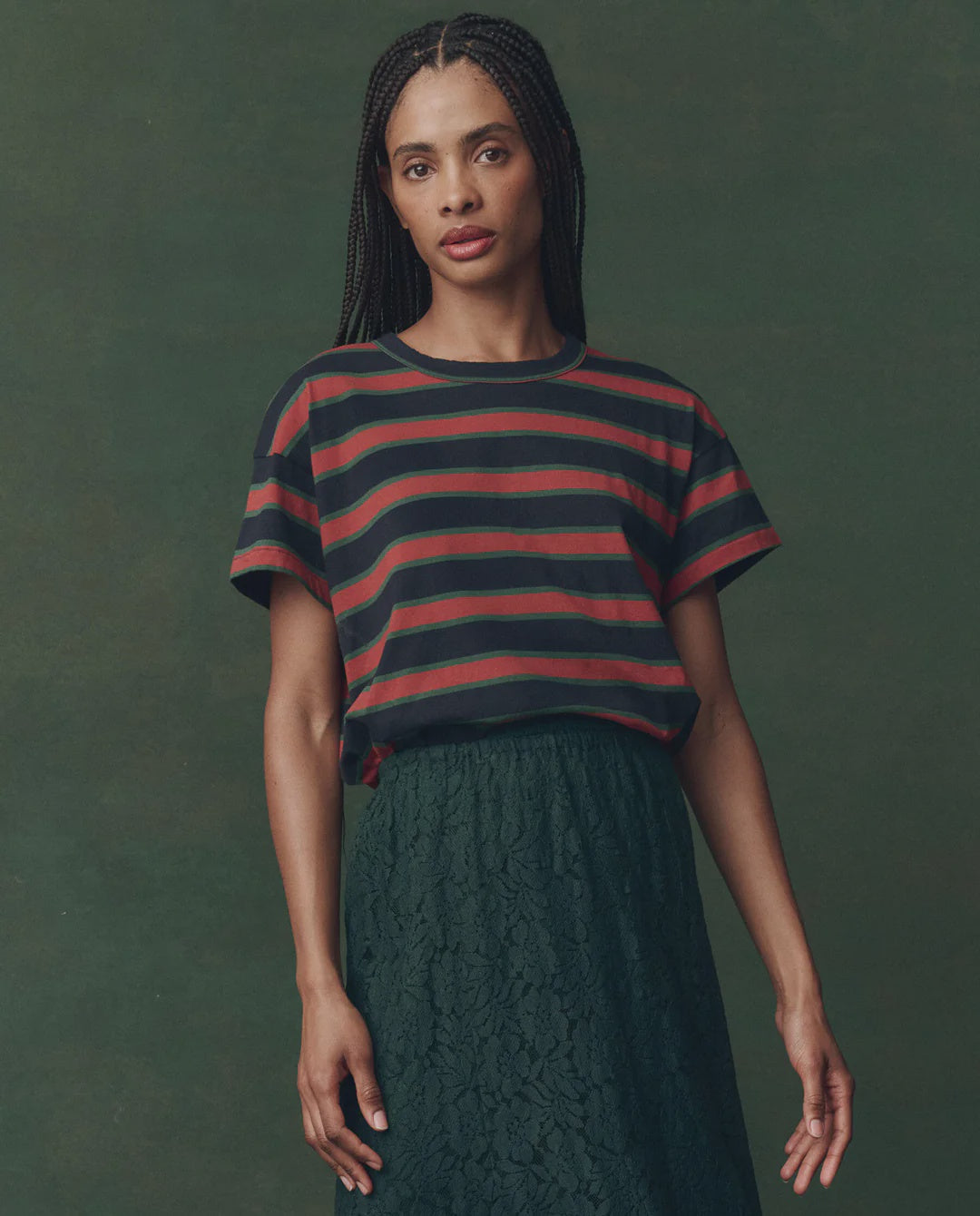 A woman with long braided hair wears The Boxy Crew F25 by The Great Inc., a 1970s-inspired vintage striped tee, paired with a high-waisted dark green lace skirt, standing against a muted green background.