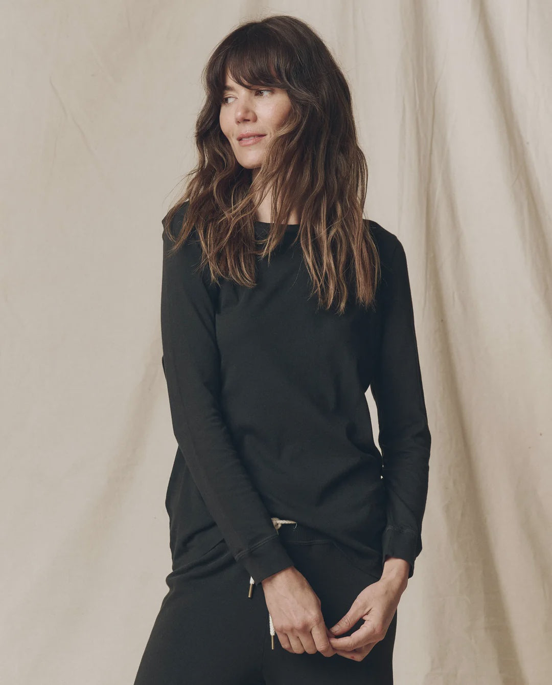 The Great Inc. Long Sleeve Slim Tee in brown, shown on a woman by a beige fabric backdrop, casual and effortless style.