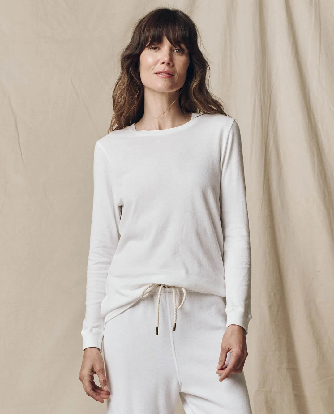 The Great Inc. Long Sleeve Slim Tee and drawstring pants in beige, worn by a smiling woman against soft backdrop.