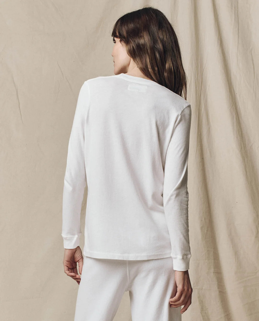 White cotton long sleeve tee by The Great Inc. worn with pants, model facing away, casual style.