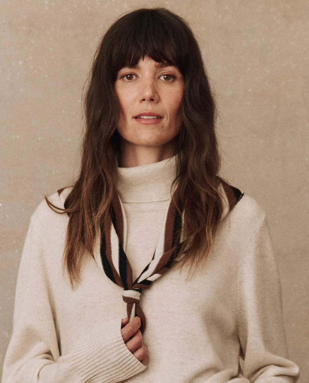 Someone with long, wavy hair and bangs is sporting The Cashmere Neckerchief by The Great Inc., paired with a light-colored turtleneck sweater. The backdrop features a neutral, textured surface.