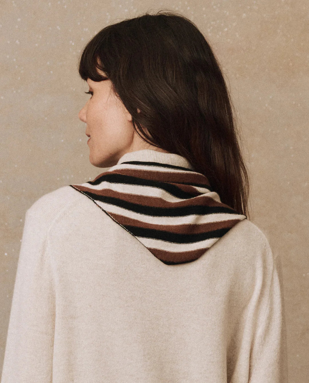 Rear view of an individual with long dark hair wearing a luxurious cream cashmere sweater, complemented by The Cashmere Neckerchief in shades of brown, black, and white draped over the shoulders. The scene is set against a speckled beige texture, showcasing the elegance of The Great Inc.