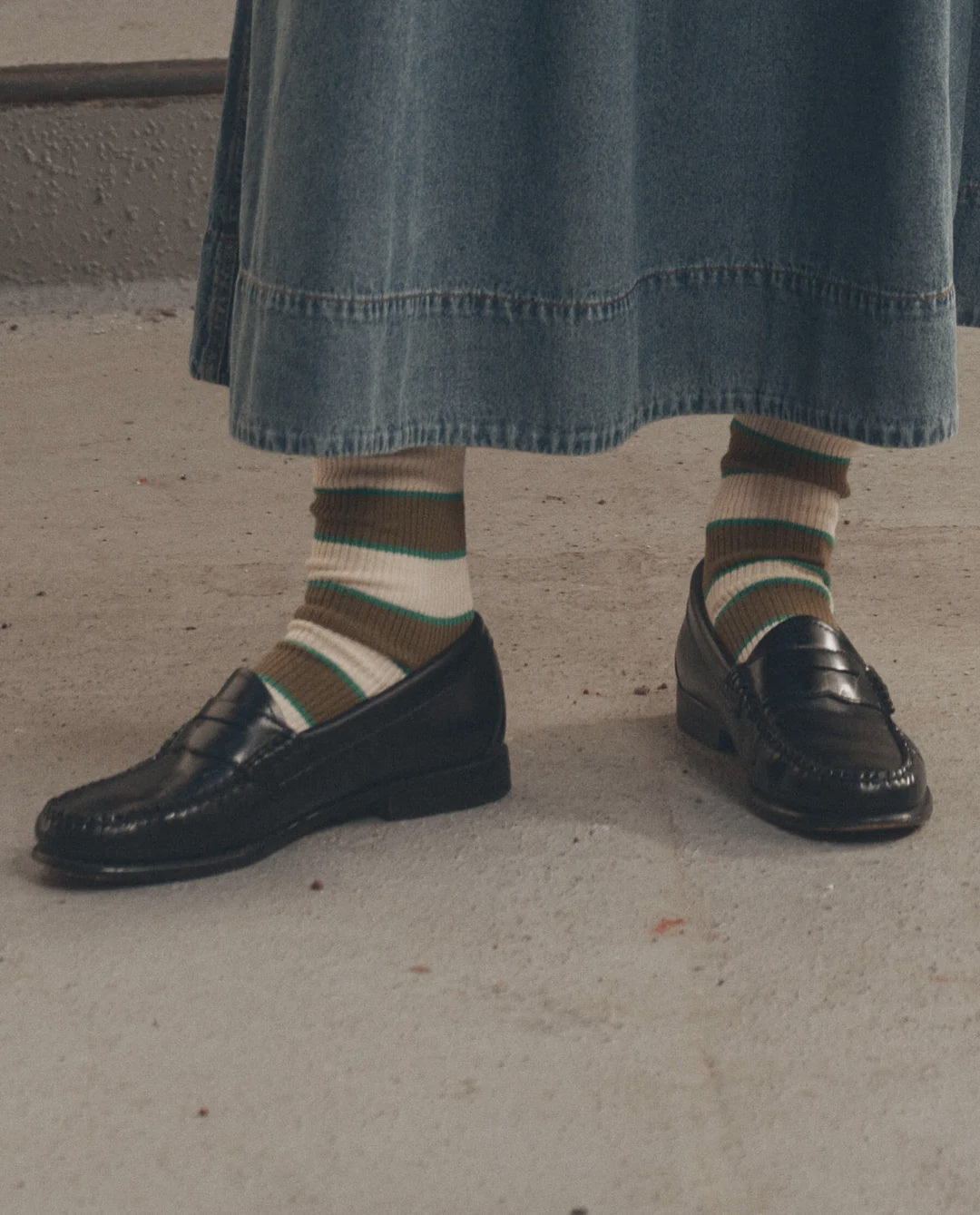 Black loafers and Collegiate Sock by The Great Inc. paired with a long denim skirt on concrete flooring.