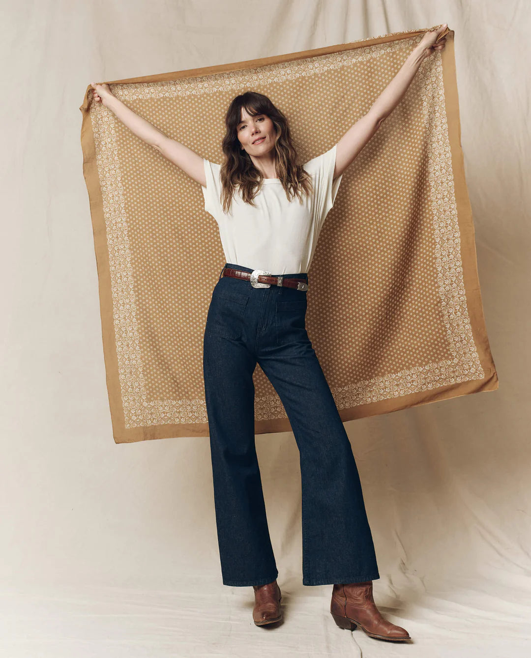 A woman smiles with arms outstretched, holding The Big Bandana by The Great Inc. behind her while wearing a white T-shirt, dark blue wide-leg jeans, and brown boots against a neutral backdrop.