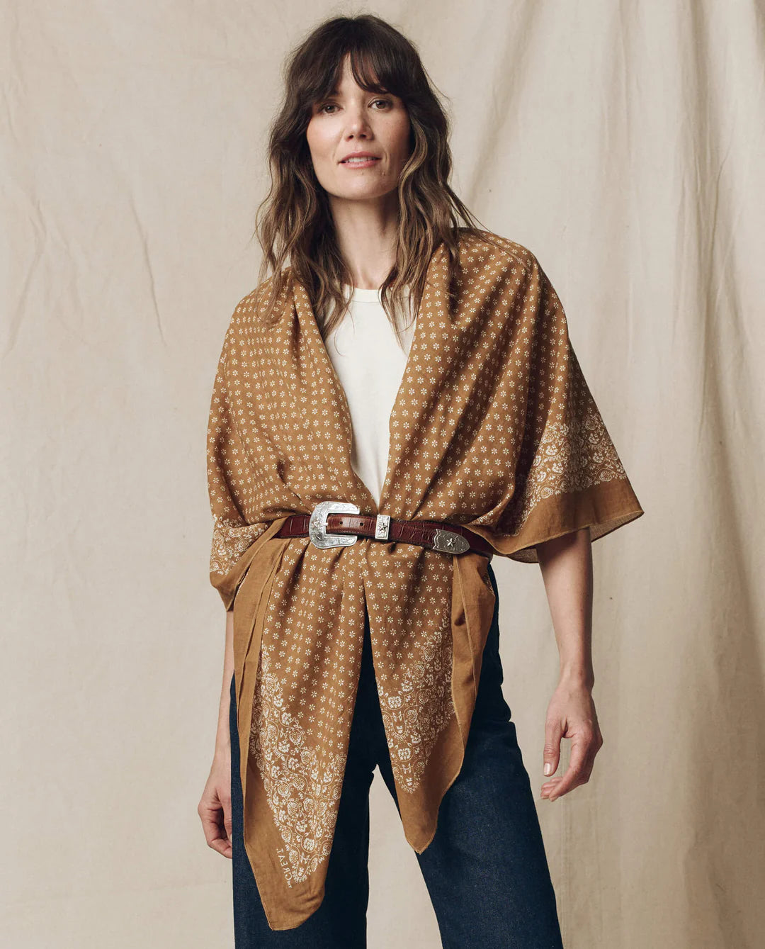 A woman with wavy brown hair and a relaxed expression wears The Great Inc.’s The Big Bandana, styled with a tan patterned shawl belted over a white top and dark jeans, standing before a neutral backdrop.