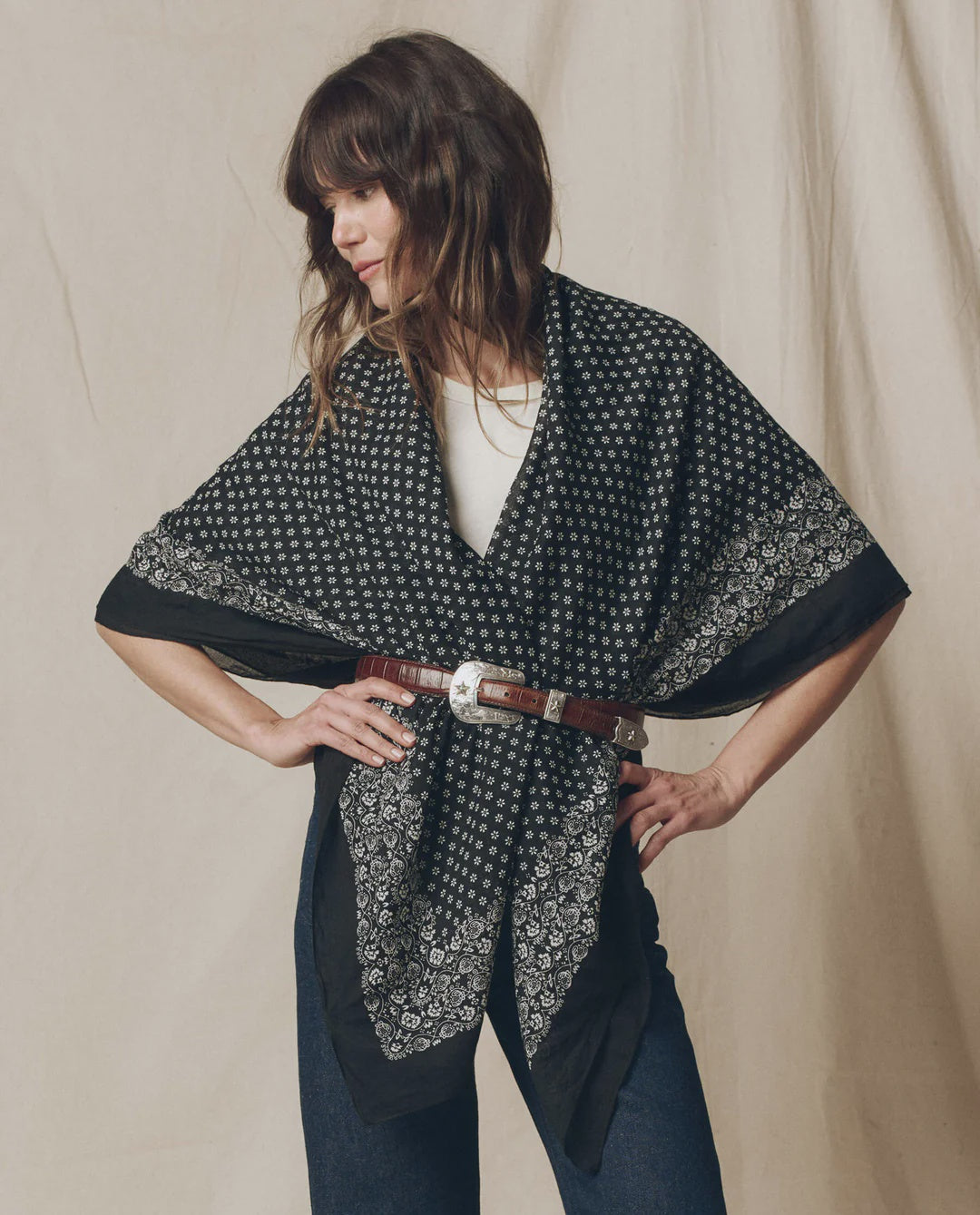 A woman stands with her hands on her hips against a neutral fabric backdrop, wearing The Big Bandana by The Great Inc. as an oversized shawl over a white top, cinched at the waist with a brown leather belt.