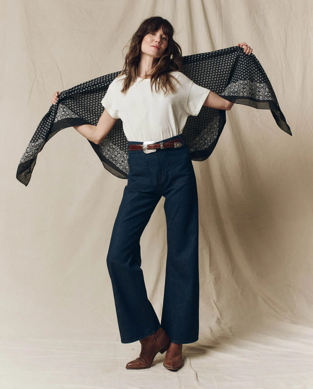 A woman stands against a beige backdrop, wearing a white tee, high-waisted dark blue wide-leg jeans, brown belt and ankle boots, holding The Big Bandana by The Great Inc. open behind her shoulders.