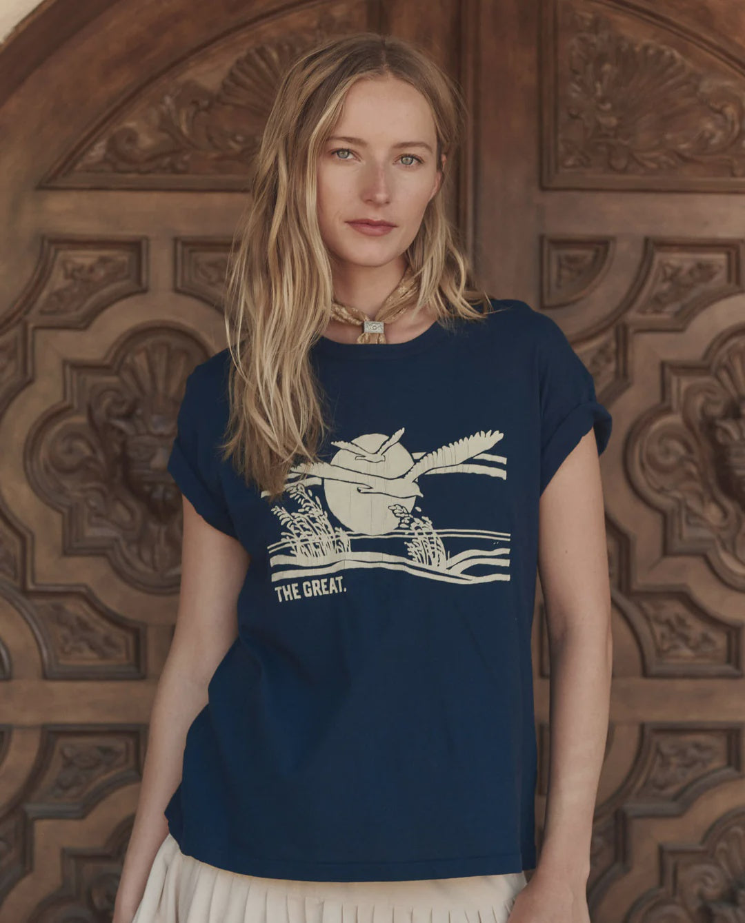A woman with long blonde hair wears The Great Inc.'s navy blue The Boxy Crew w/ Everglade Graphic tee and a cream skirt, standing by an ornate wooden door and looking at the camera—an effortlessly chic Los Angeles style.