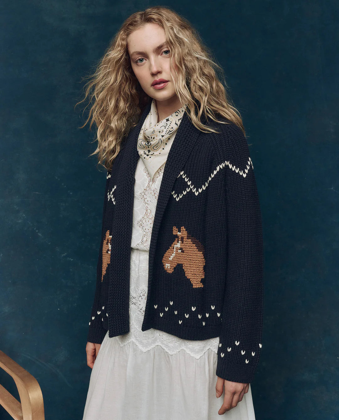 A woman with long curly blonde hair wears The Great Inc.'s Horse Lodge Cardigan, adorned with hand-embroidered horses, styled over a white lace dress and patterned scarf, set against a dark blue background evoking western nostalgia.