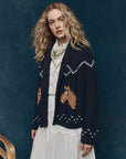 A woman with long curly blonde hair wears The Great Inc.'s Horse Lodge Cardigan, adorned with hand-embroidered horses, styled over a white lace dress and patterned scarf, set against a dark blue background evoking western nostalgia.