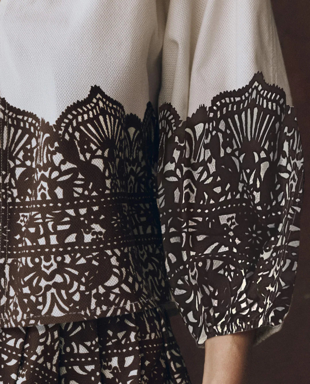 A close-up of The Chariot Top, showcasing its textured cotton with black lace patterns over white fabric and elaborate, symmetrical designs on the sleeve and bodice.