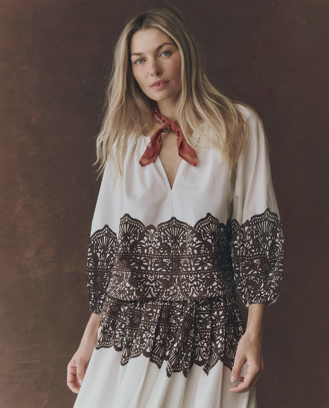 A woman with long blonde hair wears The Chariot Top—a white western lace print top—with a red scarf around her neck, standing against a brown background.