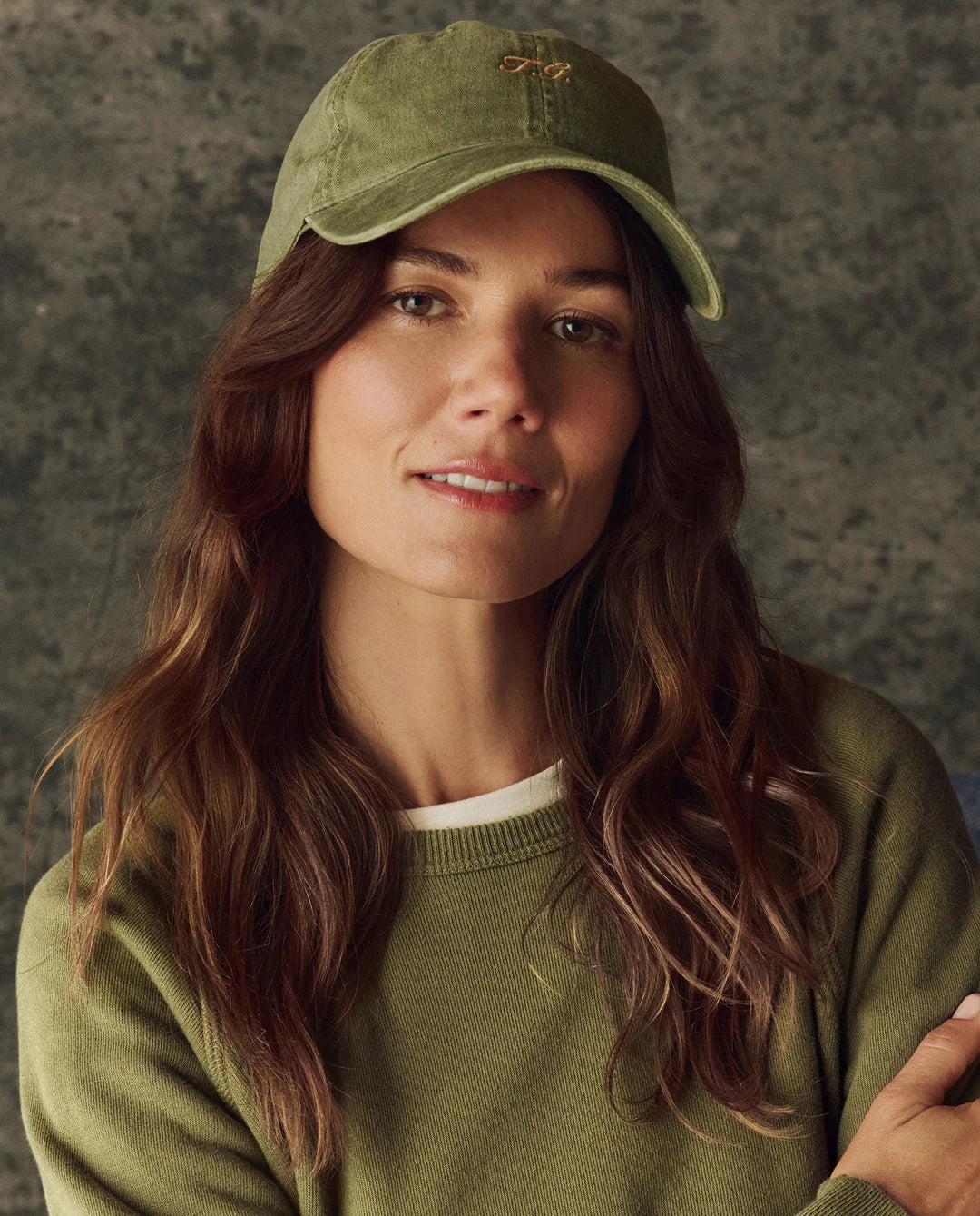 A woman with long brown hair wears The Classic Baseball Hat by The Great Inc., featuring vintage embroidery and an adjustable back, paired with a green sweater over a white shirt, standing before a neutral textured background, smiling slightly at the camera.