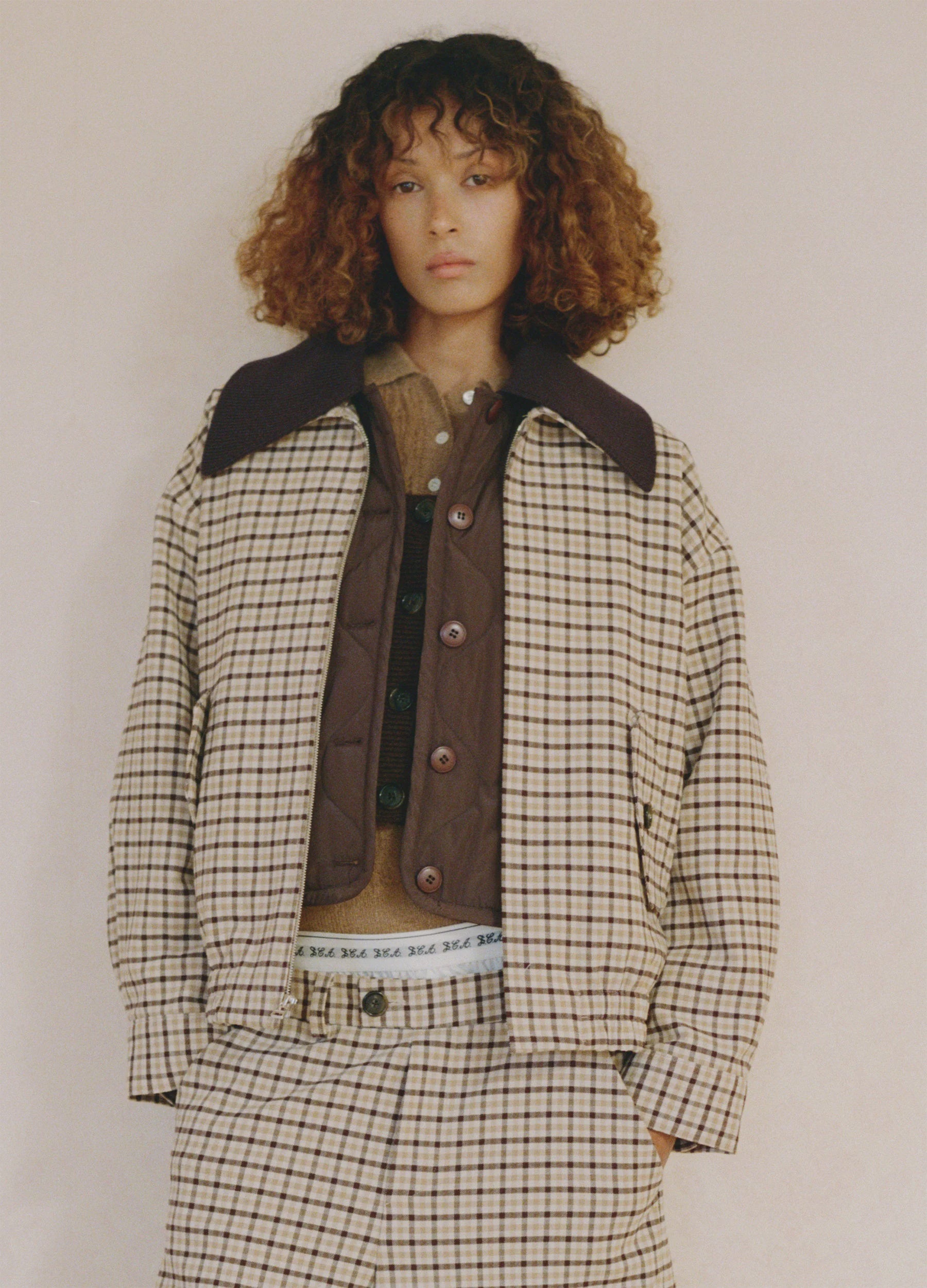 A person with curly brown hair wears the Sea New York Savannah Plaid Jacket and matching pants over a brown vest and shirt, featuring a visible waistband and relaxed fit, standing against a plain light background.