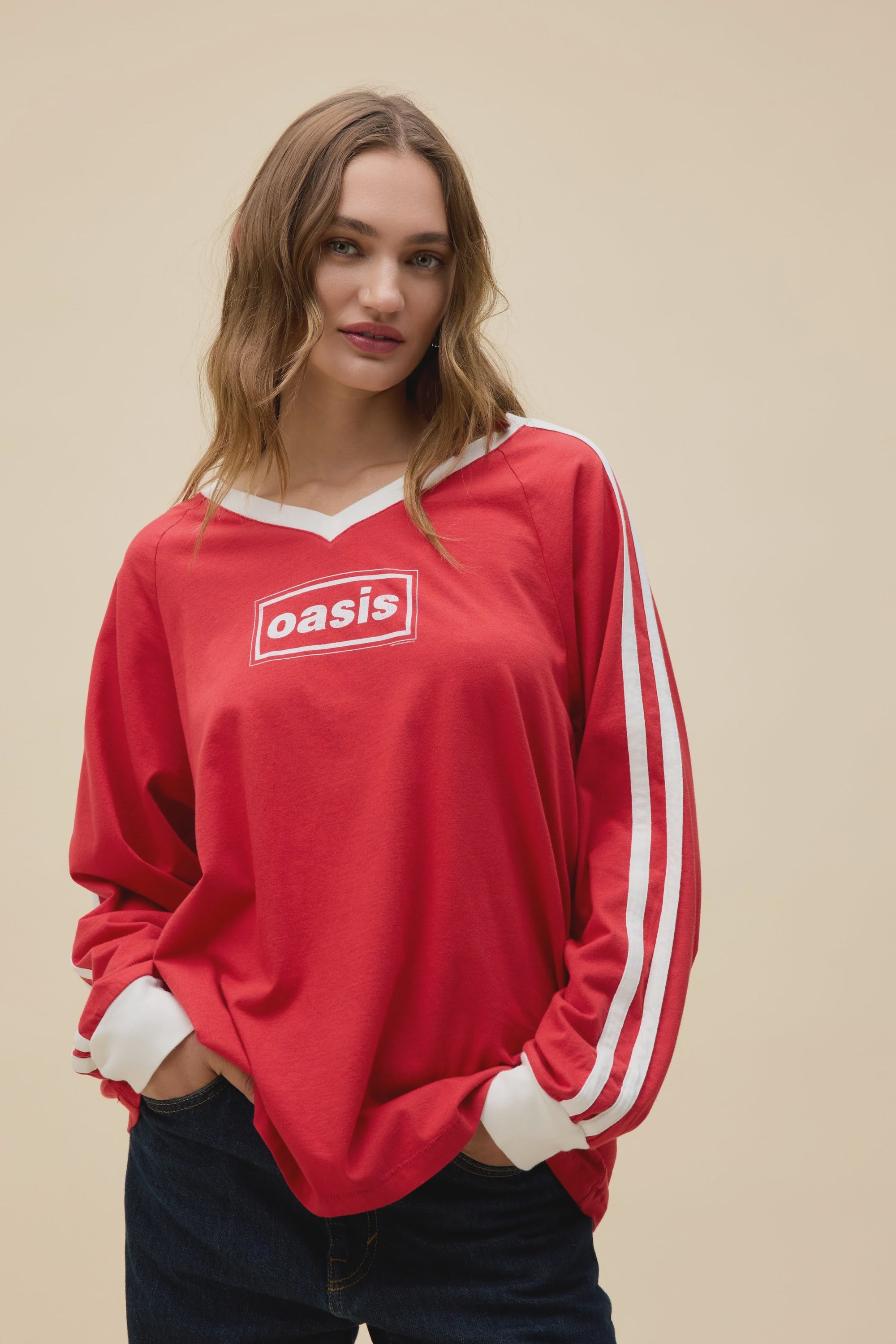 A woman with long brown hair wears the DayDreamer Oasis Box Logo red sweatshirt with white sleeve stripes, "Oasis" on the chest, and dark jeans, standing against a plain beige background.