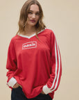 A woman with long brown hair wears the DayDreamer Oasis Box Logo red sweatshirt with white sleeve stripes, "Oasis" on the chest, and dark jeans, standing against a plain beige background.