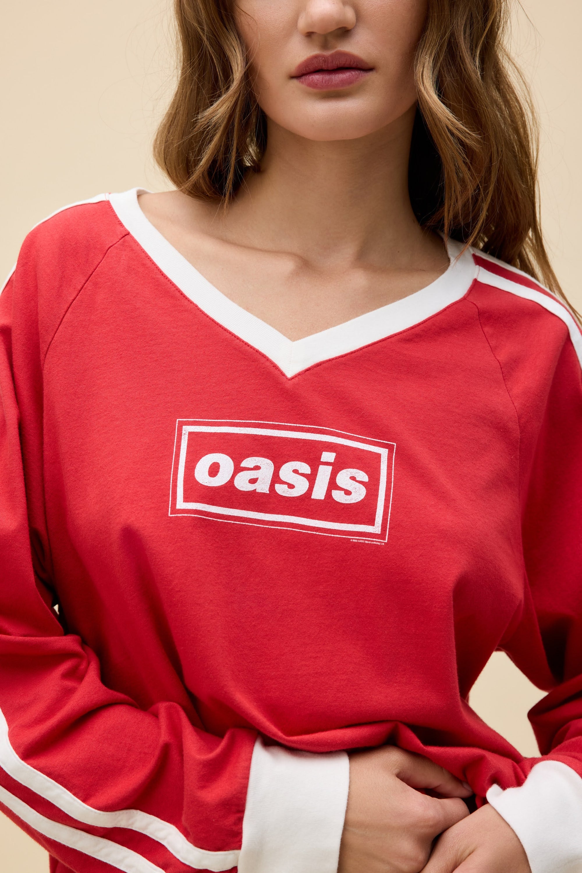 A woman with wavy brown hair wears the DayDreamer Oasis Box Logo red long-sleeve shirt featuring white trim and "Oasis" in white text inside a red box, standing against a beige background.